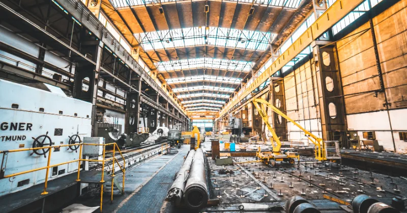 Industrial workshop interior with large machinery, steel pipes, and overhead cranes.